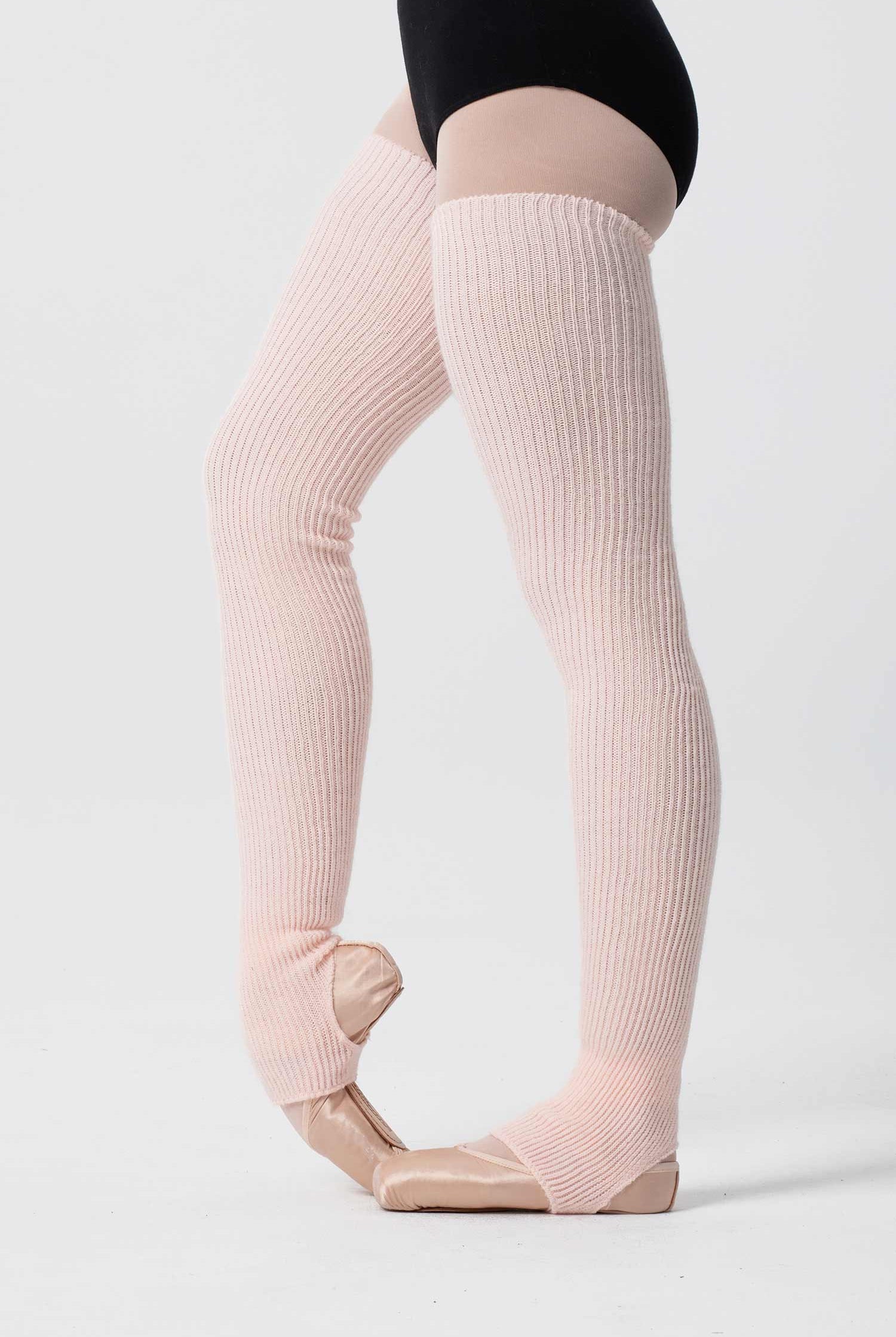Eine Person mit hellrosa Intermezzo 2657 Stulpimax flauschigen Stulpen und beigefarbenen Ballettschuhen steht auf den Zehenspitzen in einer klassischen Ballettpose vor einem weißen Hintergrund. Die Person ist in einen schwarzen Turnanzug gekleidet und der kuschelige Intermezzo Beinwärmer verleiht ihr einen zusätzlichen Hauch von Eleganz.