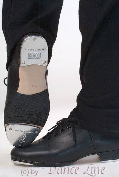 Nahaufnahme einer Person, die schwarze Steppschuhe Bloch S0301M Jazztap trägt, die die Metallkappen an den Sohlen zeigen. Ein Fuß ruht auf der Ferse und zeigt die Metallkappe darunter, während der andere flach steht. Die Person trägt eine schwarze Hose und die Schuhe sind aus hochwertigem Lederobermaterial für Langlebigkeit gefertigt.