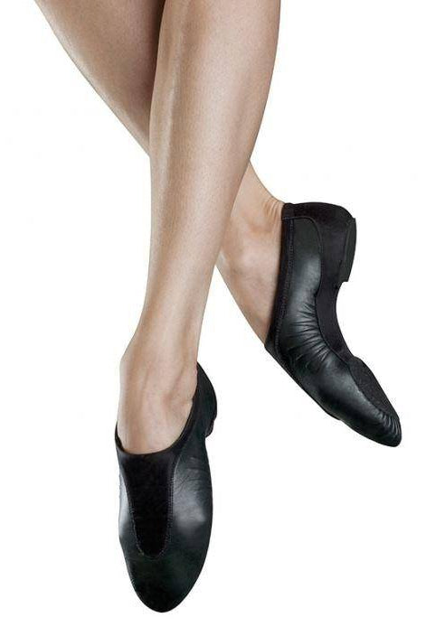 Nahaufnahme von Beinen in Jazzschuhen Bloch S0470L Pulse mit geteilter Chromledersohle, an den Knöcheln gekreuzt, vor weißem Hintergrund. Die Schuhe haben eine weiche, flexible Sohle und bieten optimalen Tragekomfort, geeignet für Tanzauftritte. Der Hautton ist hell und das Bild betont Eleganz und Schlichtheit.