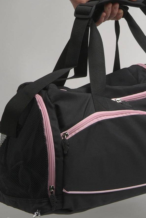 Eine Person hält eine schwarze Bloch Ballett Tasche A311 Reisetasche mit rosa Reißverschlüssen und Netzseitentaschen. Oben ist die Hand zu sehen, die die Griffe umklammert. Der Hintergrund ist in neutralem Grau gehalten. Auf der Unterseite der Tasche ist „Danceline.de“ aufgedruckt, was sie perfekt für jeden Bloch-Fan macht.
