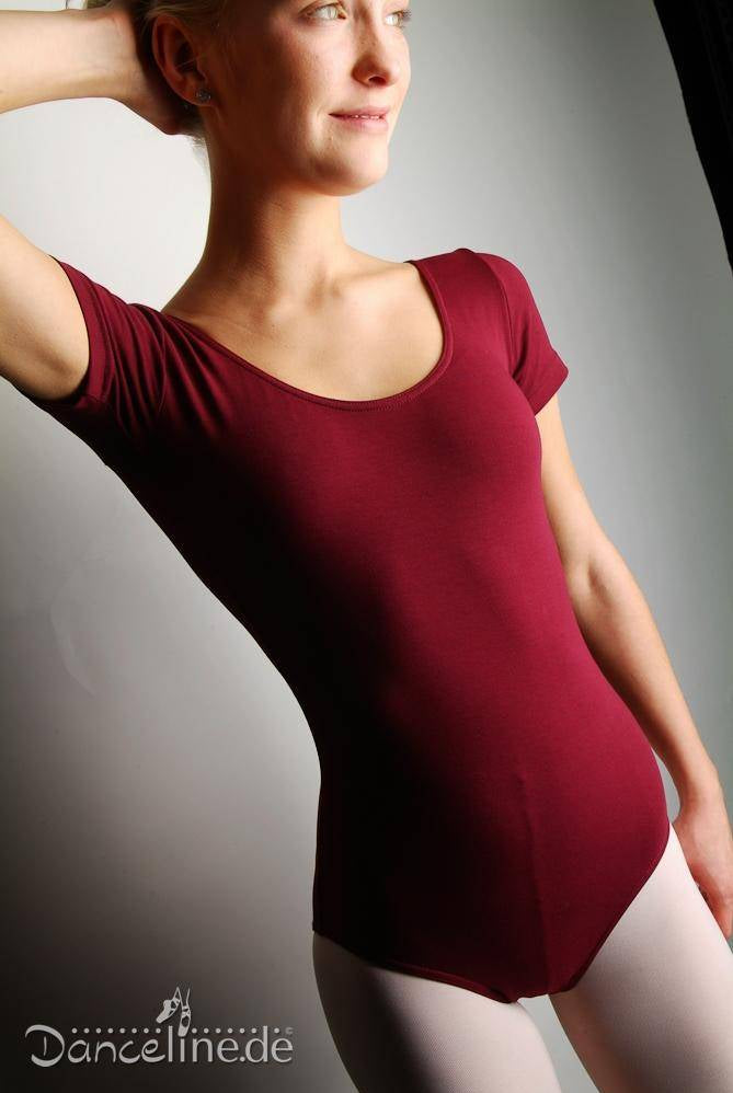 Eine Person lächelt und trägt einen kurzärmeligen, burgunderfarbenen Ballettanzug Bloch L5402 Cavalier mit weißen Strumpfhosen. Sie steht mit einem erhobenen Arm da und die andere Hand ruht auf der Hüfte. Der Hintergrund ist neutral mit sanfter Beleuchtung. In der Ecke ist das Logo „Danceline.de“ zu sehen.