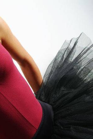 Nahaufnahme einer Balletttänzerin, die einen ärmellosen roten Turnanzug und eine schwarze Tutulette Bloch R2921 Belle von Bloch trägt. Der Oberkörper der Tänzerin und das Tutu sind deutlich zu sehen, was den Kontrast zwischen dem leuchtend roten Stoff und dem zarten schwarzen Tüll betont.
