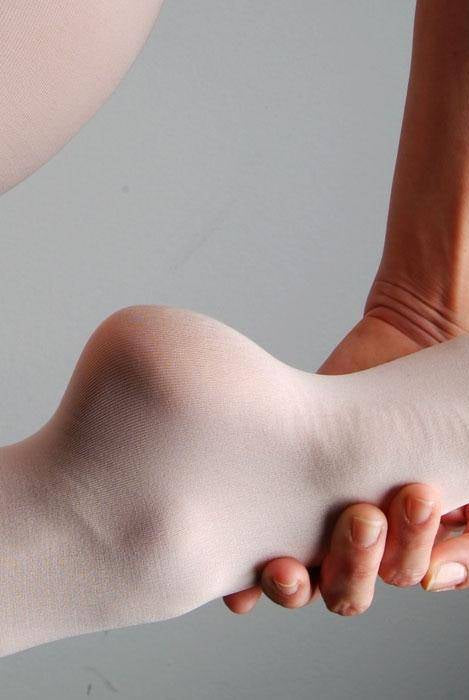 Eine Nahaufnahme des ausgestreckten und gestreckten Fußes einer Person, bekleidet mit einer weißen Ballettstrumpfhose Capezio 1818 B-Tight von Capezio. Die Hand einer anderen Person stützt die Ferse. Der Hintergrund ist schlicht hellgrau. Das Bild weist auf einer Seite ein vertikales Textwasserzeichen „Foto © www.danceline.de“ auf.