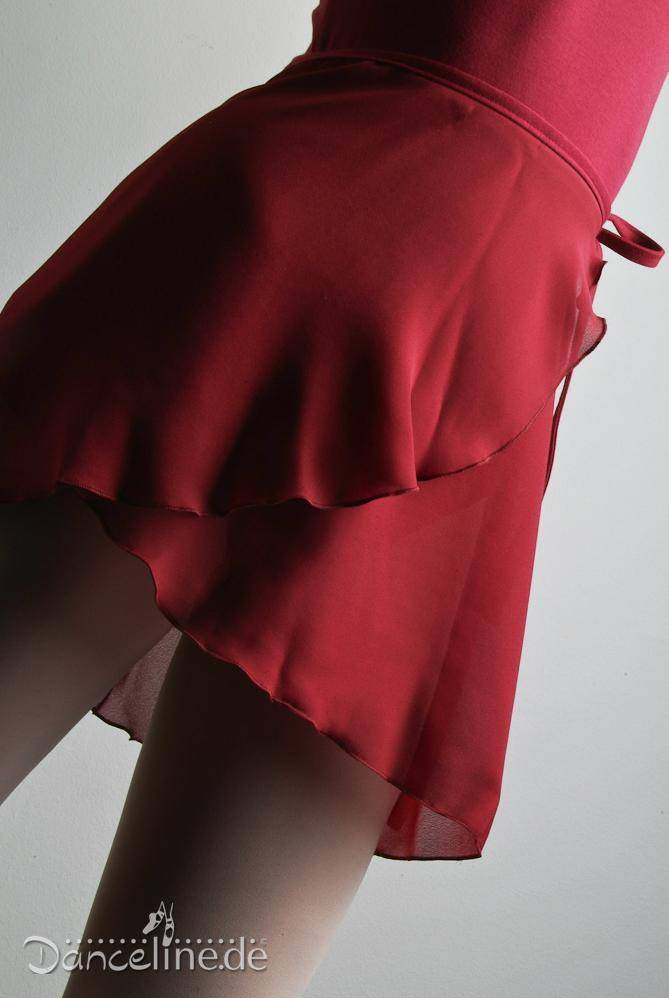 Eine Person, die den roten Wickelrock Danceries Z40 Vicky mit transparentem Überzug trägt, steht vor einem schlichten Hintergrund. Das Bild konzentriert sich auf den unteren Teil des Körpers und hebt die Textur und Bewegung des Rocks hervor, die an Ballett erinnert. In der unteren Ecke ist das Wort „Danceline.de“ sichtbar.