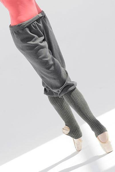 Eine Balletttänzerin in einem rosa Oberteil und grauen Hosen balanciert auf Spitze. Die Füße der Tänzerin stecken in Ballettschuhen mit Beinwärmern der Wärmehose Intermezzo 5076 Rafiness von Intermezzo und liegen auf einer weißen Oberfläche, die bei sanftem Licht einen Schatten wirft.