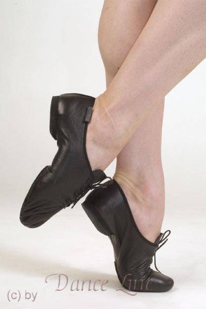 Die Unterschenkel und Füße einer Tänzerin sind in schwarzen Jazzschuhen Merlet Galion aus hochwertigem Leder zu sehen. Die Füße sind gekreuzt und ein Absatz ist angehoben. Das Design der Merlet-Schuhe, die Chromledersohle und die Flexibilität sind auf einem schlichten weißen Hintergrund zu sehen.