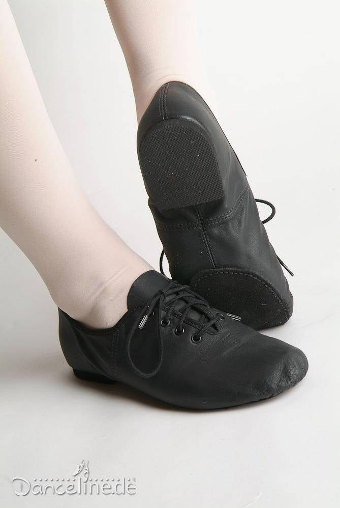 Zwei Füße sind zu sehen, die schwarze Jazzschuhe SoDanca JZE09A Jazzme Tanzschuhe mit Schnürsenkeln von SoDanca tragen. Die Person trägt außerdem hellrosa oder beige Strumpfhosen. Die Sohlen und Details der Schuhe sind sichtbar, wobei die Schuhe in einer entspannten Position zu sein scheinen.