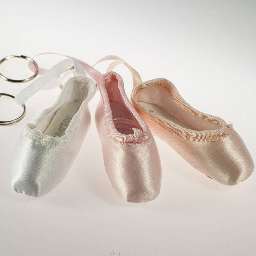 Drei Ballettschuh-Schlüsselanhänger in Pastellfarben (weiß, rosa und pfirsich) sind auf einer weißen Oberfläche abgebildet. Die **Mini-Spitzenschuh-Anhänger SoDanca KCE01 Minipoint**-Schlüsselanhänger sind nebeneinander angeordnet, jeder mit einem daran befestigten Schlüsselring. Unten steht ein Logo und der Text „Danceline.de Fachversand für Ballettartikel“. Ein perfektes Geschenk für Ballettliebhaber!