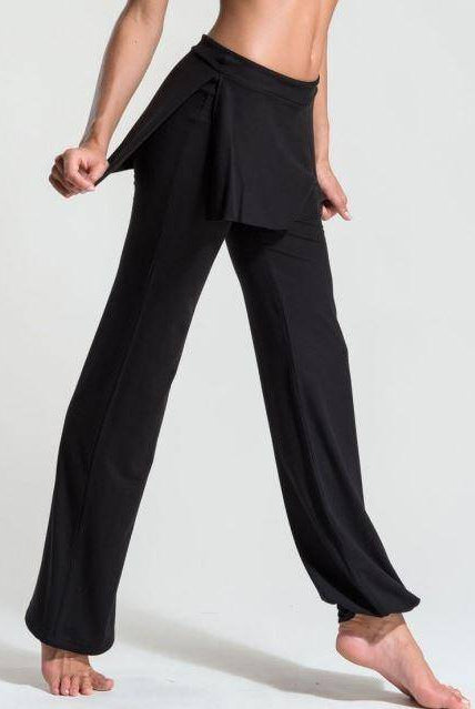 Eine Person trägt die Hose TempsDanse Zest in Schwarz von Temps Danse. Das Foto zeigt nur die untere Hälfte des Körpers, während sie auf einem Bein steht und das andere Bein leicht angewinkelt ist, wodurch der fließende Viskosestoff und die Wickelauflage an den Hüften sichtbar sind.
