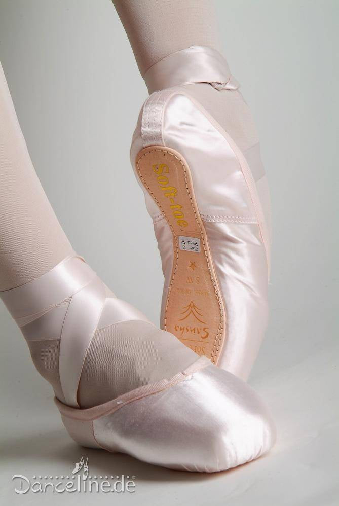 Das Bild zeigt eine Nahaufnahme der Füße einer Balletttänzerin in Softpoints Sansha DP802, die anmutig posiert. Die Schuhe sind aus hellrosa Satin mit Bändern um die Knöchel. Das Wort „Sansha“ und ein Text sind auf der Sohle eines Schuhs zu sehen, was darauf hinweist, dass sie ideal für fortgeschrittene Schüler sind.