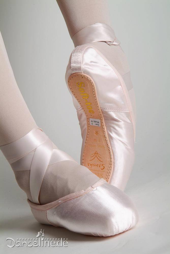 Das Bild zeigt eine Nahaufnahme der Füße einer Balletttänzerin in Softpoints Sansha DP802, die anmutig posiert. Die Schuhe sind aus hellrosa Satin mit Bändern um die Knöchel. Das Wort „Sansha“ und ein Text sind auf der Sohle eines Schuhs zu sehen, was darauf hinweist, dass sie ideal für fortgeschrittene Schüler sind.