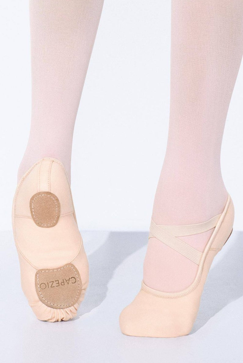 Nahaufnahme der Unterschenkel und Füße einer Tänzerin, die hellrosa Ballettschuhe Capezio 2037W Hanami und Strumpfhosen trägt. Der linke Fuß steht flach auf dem Boden und zeigt die geteilte Chromledersohle mit der Aufschrift „Capezio“, während der rechte Fuß leicht angehoben ist und elastische Bi-Stretchleinen-Riemen zeigt.