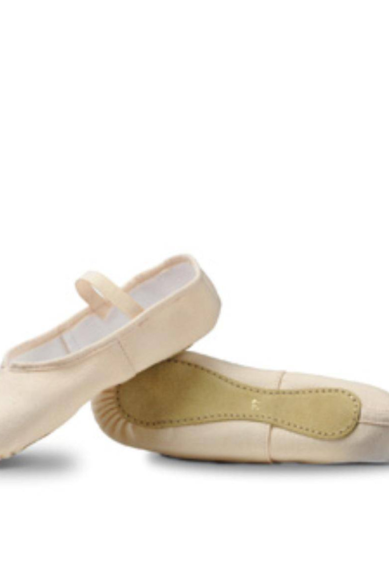 Ein Paar beige Ballettschuhe Merlet Envol mit leichtem Glanz, perfekt für Kinder oder Anfänger. Ein Schuh liegt flach auf der Oberfläche, während der andere darauf balanciert, wodurch die Struktur der Unterseite und das Sohlendesign sichtbar werden. Der flache Schuh verfügt über einen elastischen Riemen an der Oberseite für einen sicheren Sitz.