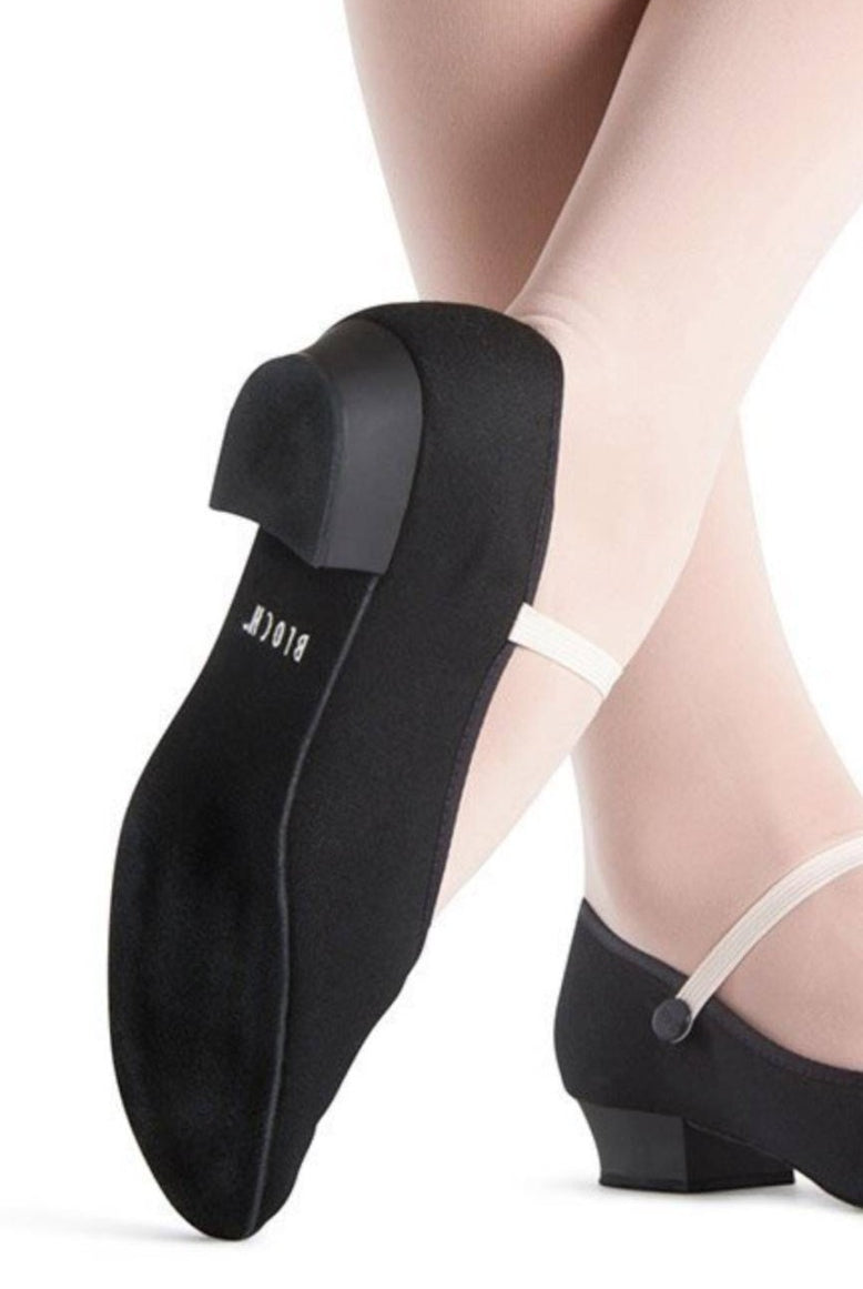 Nahaufnahme der Beine einer Tänzerin, die hellrosa Strumpfhosen und schwarze Charakterschuhe Bloch S0326 Accent mit niedrigem Absatz und weißem Gummiband trägt. Der Markenname „Bloch“ ist auf der Sohle des angehobenen Fußes sichtbar und hebt die gepolsterte Einlegesohle hervor.