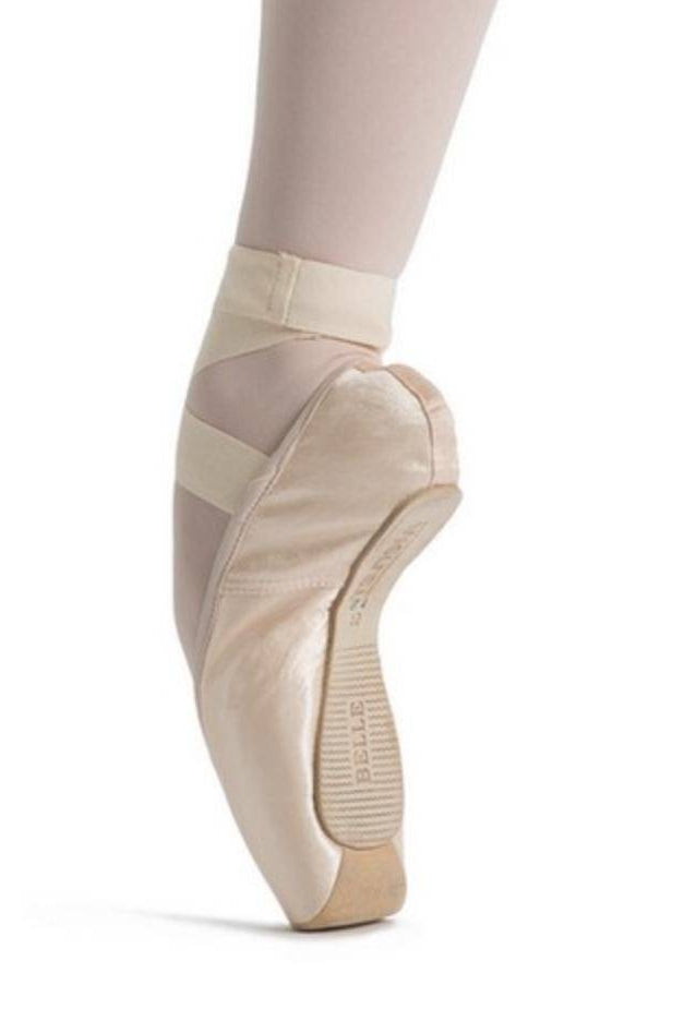 Nahaufnahme des Fußes einer Balletttänzerin auf Spitze, die rosa Spitzenschuhe Merlet Belle mit gekreuzten elastischen Riemen über hellen Strumpfhosen trägt. Die Ferse der Anfängerin ist angehoben, wodurch die Kurven und die Struktur des Merlet-Spitzenschuhs und die Kraft des Fußes hervorgehoben werden.