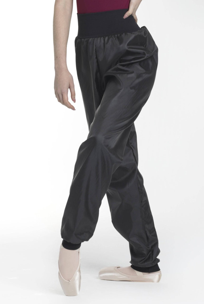 Eine Person in einem roten Oberteil, einer schwarzen hoch taillierten Intermezzo Saunahose Pana 5256 und rosa Ballettschuhen posiert vor einem schlichten weißen Hintergrund. Die locker sitzende Hose mit glänzender Textur bietet optimale Bewegungsfreiheit, da die Person mit einem leicht gebeugten Bein steht.