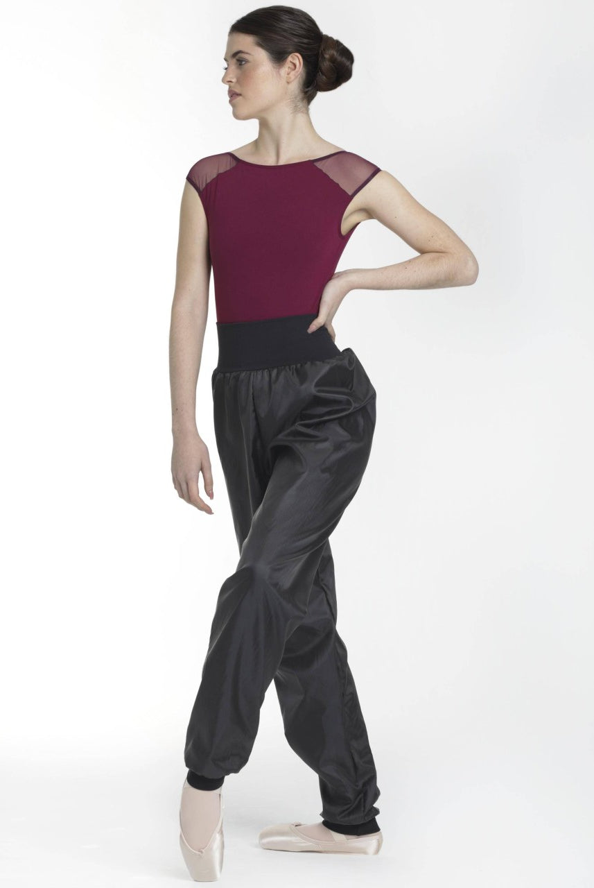 Eine Person steht in einer Ballettpose und trägt einen kastanienbraunen Leotard mit transparenten Schulterteilen, die hoch taillierte schwarze Intermezzo Saunahose Pana 5256, die Bewegungsfreiheit bietet, und Ballettschuhe. Die Person hat ihr Haar zu einem glatten Knoten gestylt und blickt leicht zur Seite, während ein Arm auf der Hüfte und der andere an der Seite ruht.