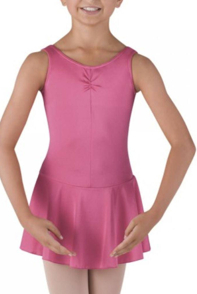 Eine junge Person in einem rosa Kinder Ballettanzug Bloch BU902C mit angenähtem Rock steht mit gerundeten Armen auf Taillenhöhe und trägt beige Strumpfhosen. Der Hintergrund ist schlicht weiß.