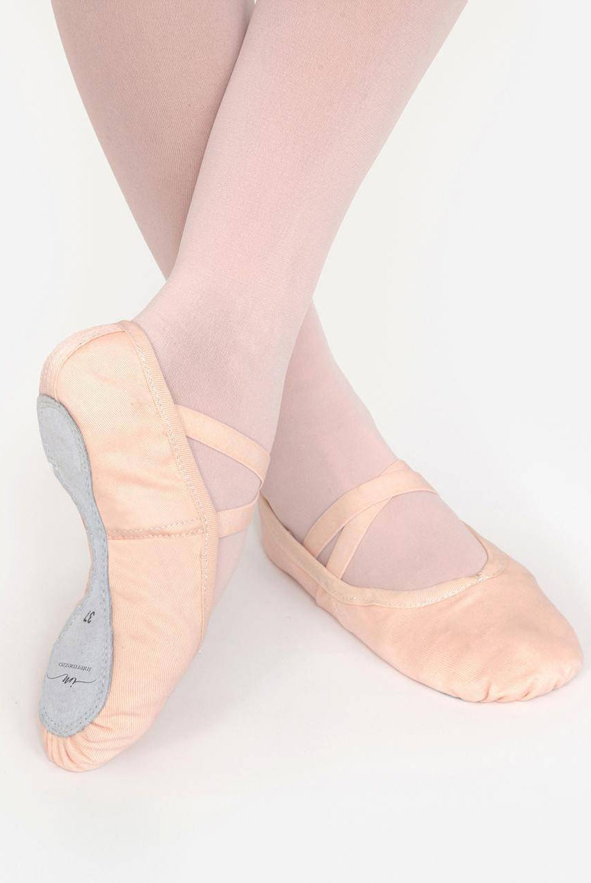 Eine Nahaufnahme der Unterschenkel und Füße einer Person, die rosa Intermezzo Ballettschuhe Intermezzo 7254 mit Chromledersohle und hellrosa Strumpfhosen trägt. Die Füße sind zart übereinandergeschlagen und zeigen die Ballettschuhe mit weichen, runden Zehen und elastischen Riemen darüber. Der Hintergrund ist schlicht weiß.