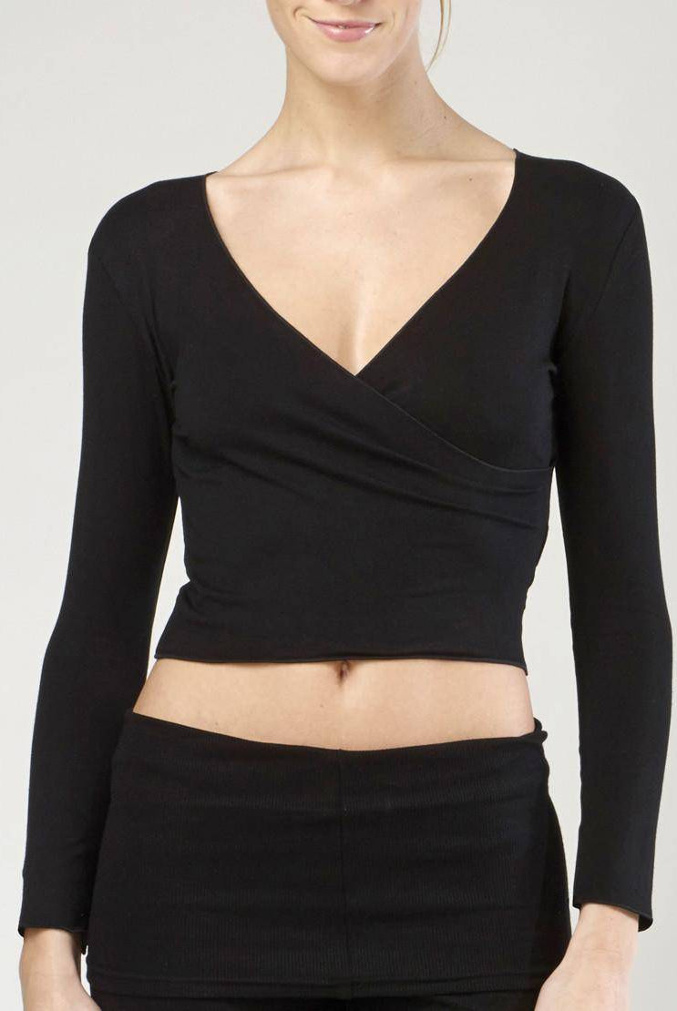 Eine Person trägt das stylische Wickeltop TempsDanse Vidy von Temps Danse. Das schwarze, langärmlige V-Ausschnitt-Crop-Top hat ein Wickeldesign, das an eine traditionelle Wickeljacke erinnert. Das Oberteil ist mit einer passenden schwarzen Hose kombiniert. Das Gesicht der Person ist teilweise sichtbar, mit einem subtilen Lächeln vor einem schlichten und hellen Hintergrund.
