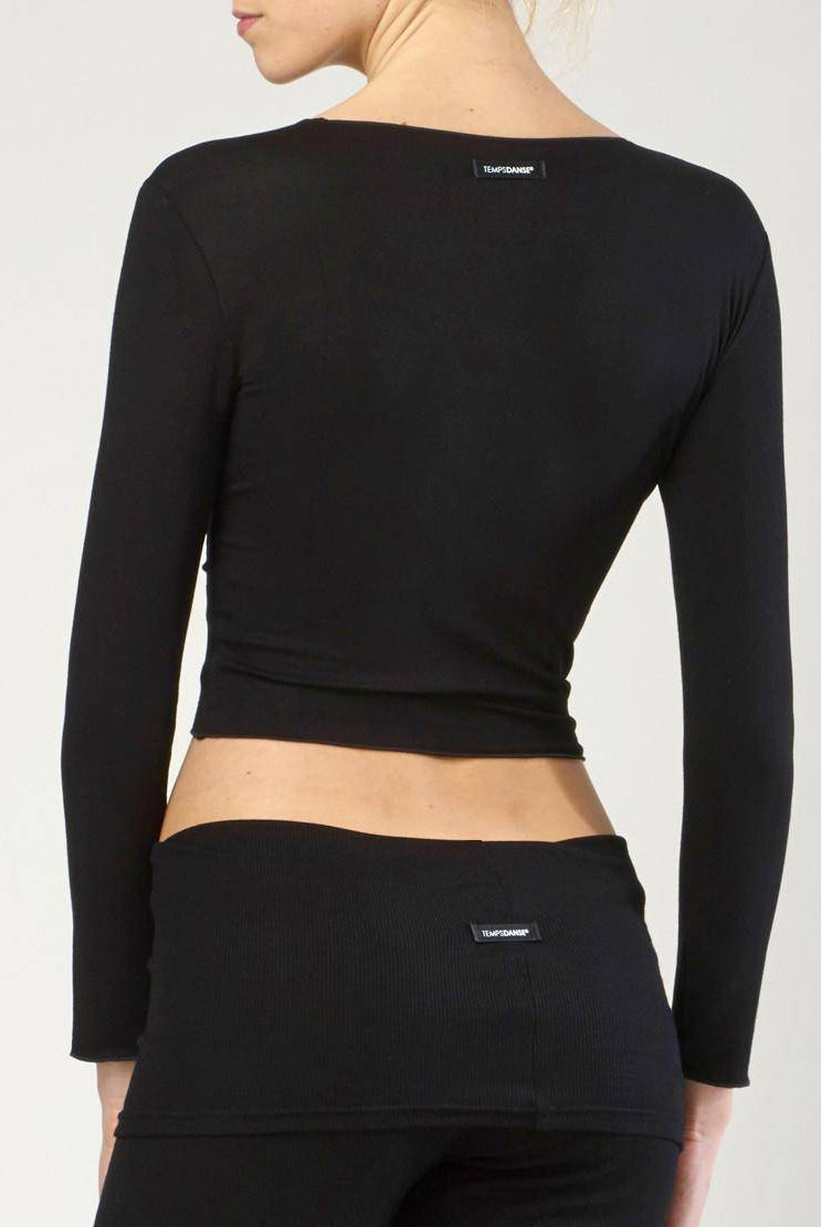 Eine Frau mit blonden Haaren trägt ein schwarzes Temps Danse Wickeltop Vidy und eine schwarze Hose. Beide Kleidungsstücke haben kleine schwarze Etiketten mit weißem Text darauf. Sie steht vor einem neutralen Hintergrund.