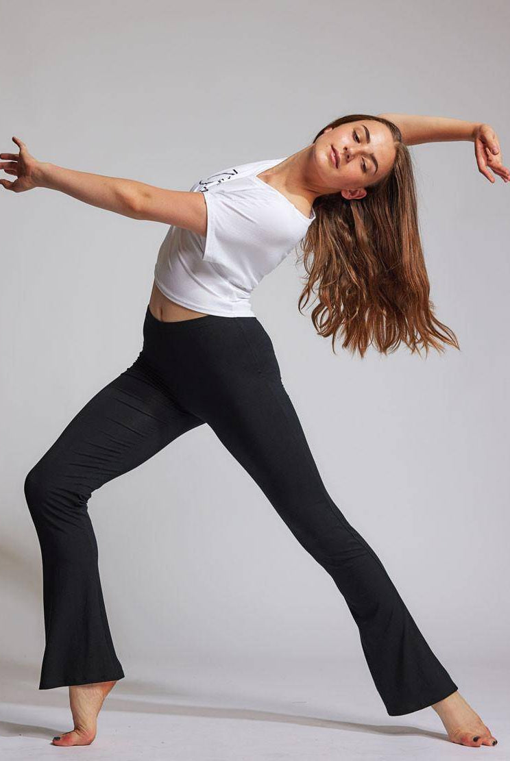 In einem Studio führt Luna eine Tanzbewegung aus, ein Bein ist nach hinten ausgestreckt und das andere leicht gebeugt. Ihr langes braunes Haar weht, während sie auf ihren Zehenspitzen steht und die schwarze Jazzhose TempsDanse Luna und ein weißes bauchfreies Top trägt. Mit anmutig ausgestreckten Armen verkörpert sie die Essenz von Temps Danse.