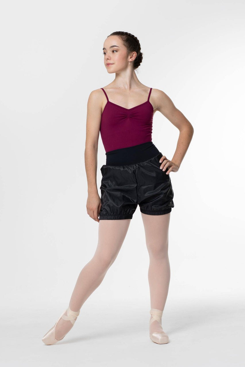 Eine Ballerina in einem kastanienbraunen Trikot, schwarzen Wärmeshorts Intermezzo 5298 Panadel und rosa Strumpfhosen steht in einer Tanzpose mit einem Bein nach vorne. Ihre Hände sind auf ihren Hüften und sie trägt Spitzenschuhe. Ihr Haar ist ordentlich zu einem geflochtenen Knoten gestylt. Der Hintergrund ist schlicht weiß.