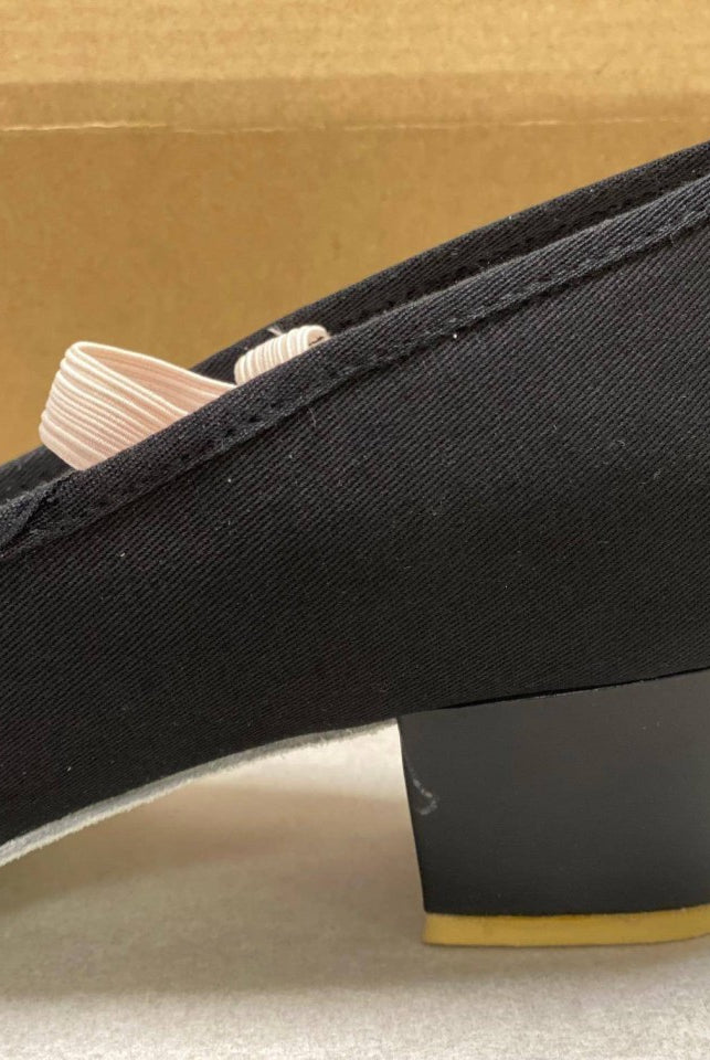 Eine Nahaufnahme eines einzelnen schwarzen Danceries Charakterschuhs Z66, der auf einer hellen Oberfläche mit einer braunen Schachtel im Hintergrund liegt. Der Schuh, ideal für Schüler der Royal Academy of Dance, verfügt über eine dicke Sohle und ein dezentes Banddetail in der Nähe der Ferse.