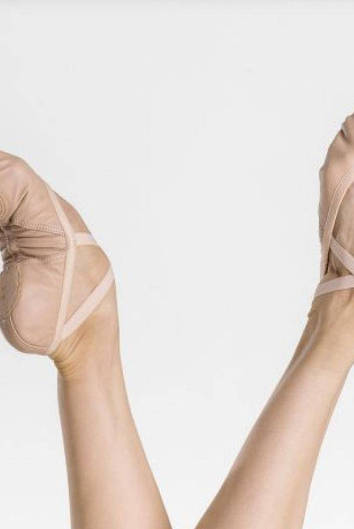 Ein Paar Füße in hellrosa Ballettschuhen WearMoi Pluton von Dance Line mit gekreuzten elastischen Riemen ragen vor einem weißen Hintergrund in die Luft. Die gut ausgeprägten Muskeln und die ausgeglichene Haltung der Füße betonen die typische Anmut und Kraft einer Balletttänzerin.