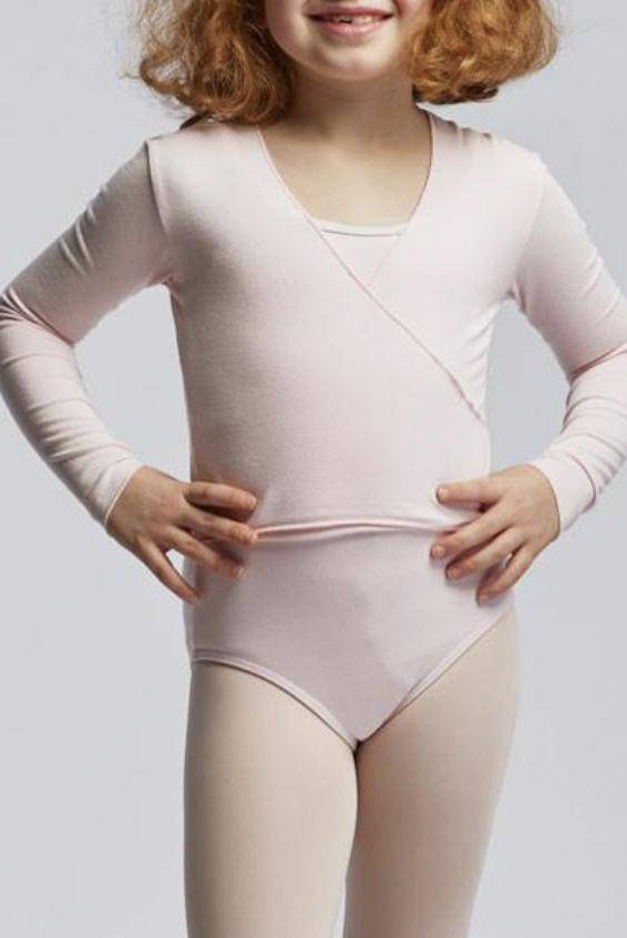 Ein kleines Kind mit lockigem Haar steht vor einem schlichten Hintergrund. Es trägt ein hellrosa Ballett-Outfit, darunter das Temps Danse Wickeltop TempsDanse Vidy Junior aus weicher Viskose, das die Muskeln warm hält. Die Hände ruhen auf den Hüften und es hat ein kleines Lächeln im Gesicht.