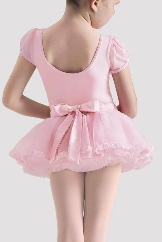 Ein kleines Kind trägt eine Kindertütülette Bloch CR4061Rose von Bloch, mit kurzen Ärmeln und einem Tüll-Tutu, von hinten betrachtet. Das Tutu weist Rüschenlagen auf und ist an der Taille mit einer großen Satinschleife verziert. Das Kind hat kurze Haare und steht vor einem schlichten, hellgrauen Hintergrund.