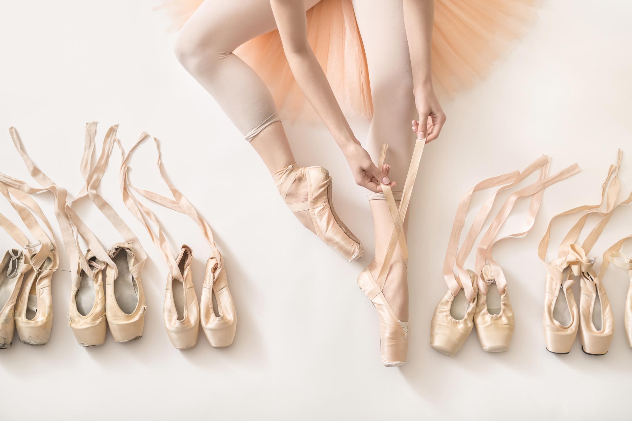 Viele Ballettschuhe in gebrauchten Zustand mit Bändern. In der Mitte sind die Beine einer Ballerina, die einen Spitzenschuh fertig anhat und den anderen gerade bindet.