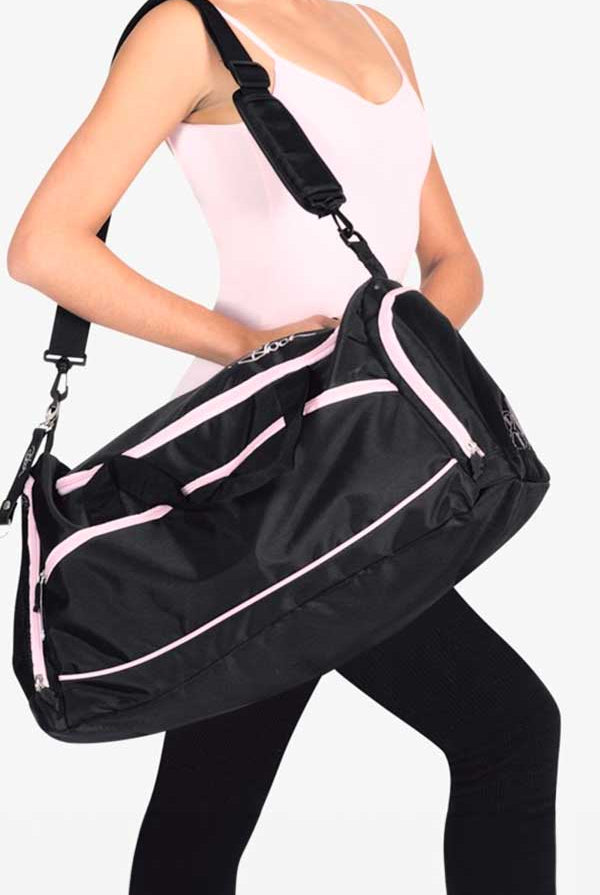 Eine Person in einem rosa Tanktop und einer schwarzen Hose trägt eine große Bloch Ballett Tasche A311 Duffle mit rosa Reißverschlüssen und Akzenten. Die Tasche wird mit dem Schultergurt über die Schulter gehängt. Der Hintergrund ist schlicht weiß.