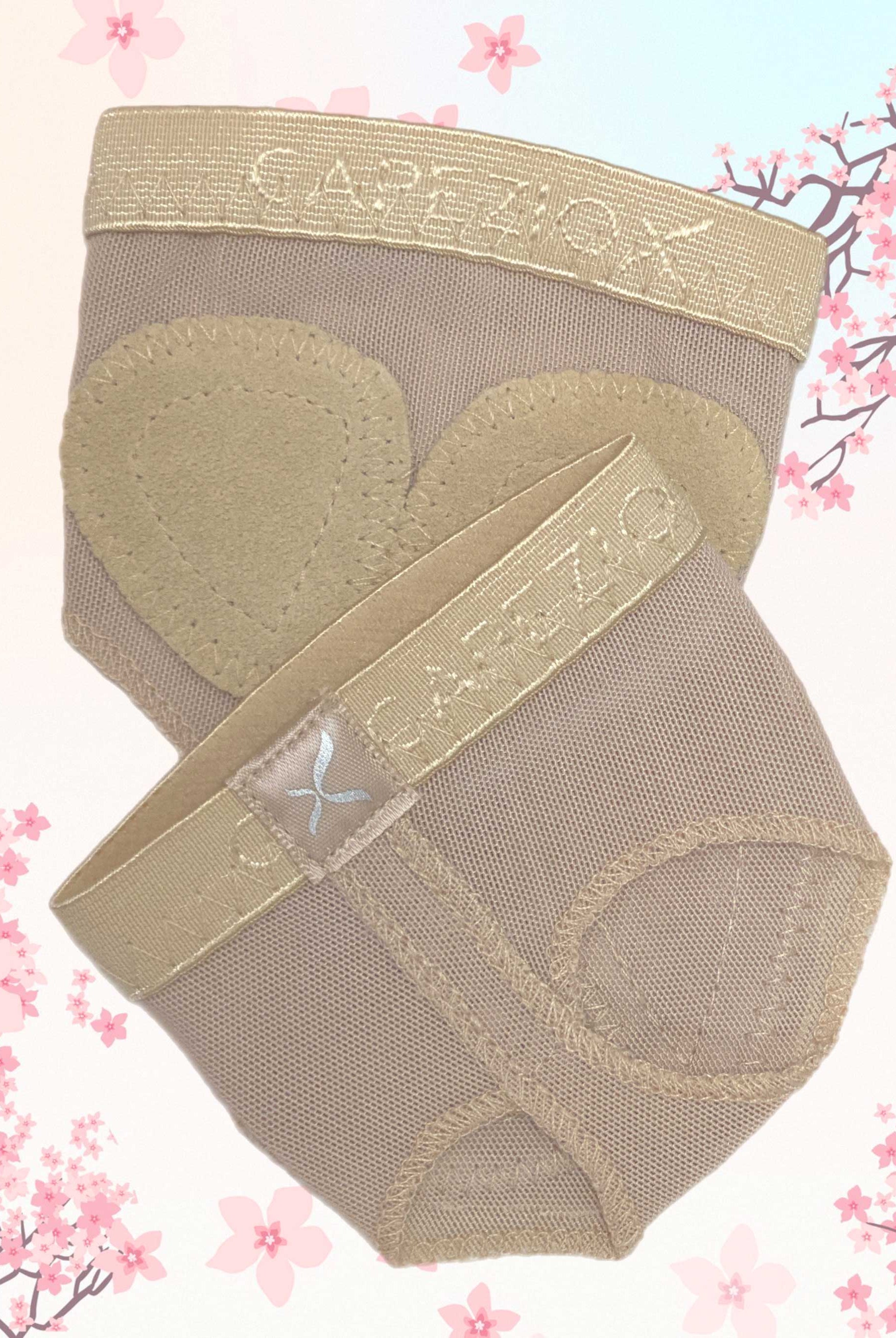 Ein Paar Ballenschutz Capezio H07B FootUndeez in Beige, mit herzförmigen Polstern aus Spandex-Material vor einem Hintergrund aus hellrosa Kirschblüten-Illustrationen. Ausgestattet mit einem elastischen Riemen ist der Markenname „Capezio“ sowohl auf dem Bund als auch auf dem Riemen deutlich zu erkennen.