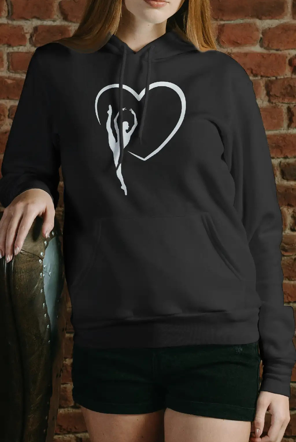 Eine Person steht vor einer Backsteinmauer und trägt den Dance Line Ballett Hoodie aus der Danceline High Legs Kollektion, der ein Herzdesign mit einer stilisierten Figur darin aufweist. Sie trägt außerdem schwarze Shorts und stützt eine Hand auf einen Sessel.