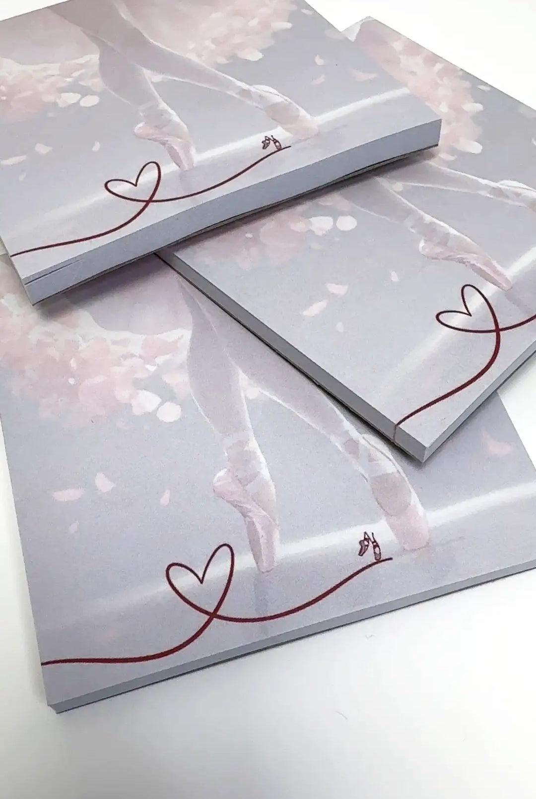 Piles of Block Danceline-Notizbücher „Dancing around“ von Dance Line mit einem Ballettthema in sanften Rosa- und Weißtönen. Das Coverdesign hebt Ballettschuhe und eine durchgehende rote Linie hervor, die in der Ecke eine Herzform bildet.