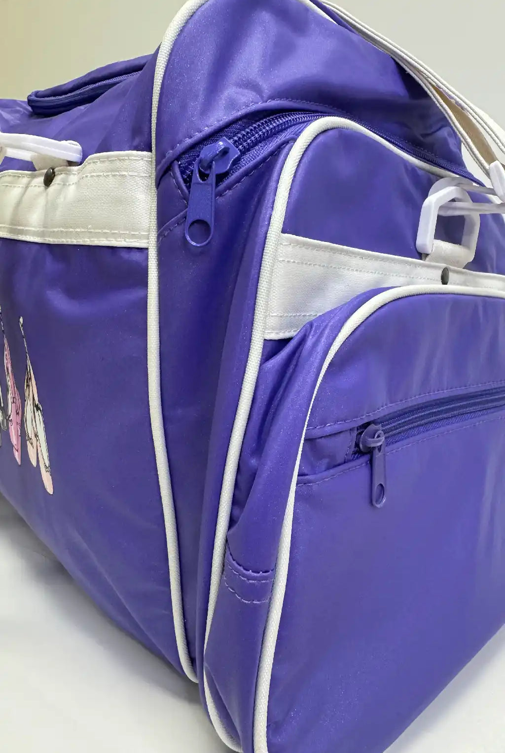 Eine Nahaufnahme der Balletttasche GD177 von Dance Line mit violetter Außenseite mit weißen Reißverschlüssen und Verzierungen. Diese Sporttasche verfügt über mehrere Fächer, darunter eine Seitentasche, und zeigt auf der linken Seite ein Design.