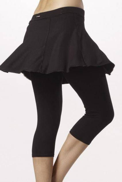 Person, die nur ihren Unterkörper zeigt und einen schwarzen Skaterrock über der bequemen, elastischen Viskose-Caprihose Temps Danse Hose TempsDanse Zazou trägt. Der Rock ist am Saum ausgestellt und die Leggings sind knapp unter dem Knie abgeschnitten. Der Hintergrund ist schlicht und hell.