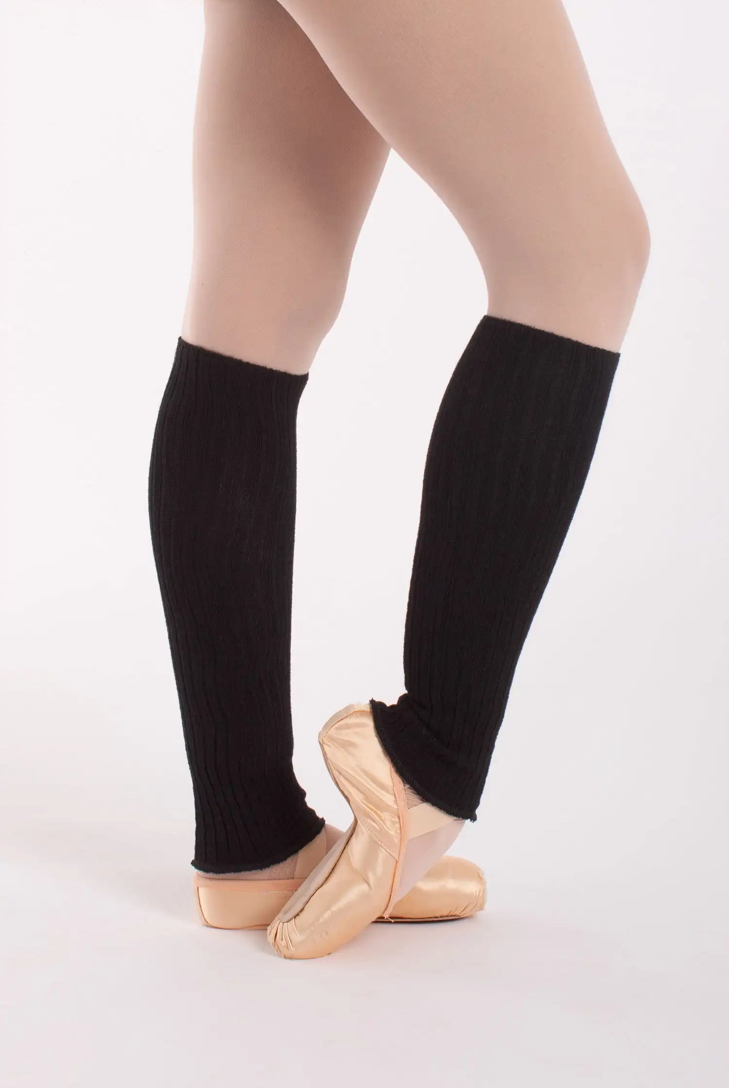 Eine Ballerina steht auf Spitze auf einem weißen Hintergrund und trägt pfirsichfarbene Ballettschuhe, beige Strumpfhosen und die schwarzen Beinwärmer „Stulpen 50 cm Intermezzo 2034 Danae“ von Intermezzo.