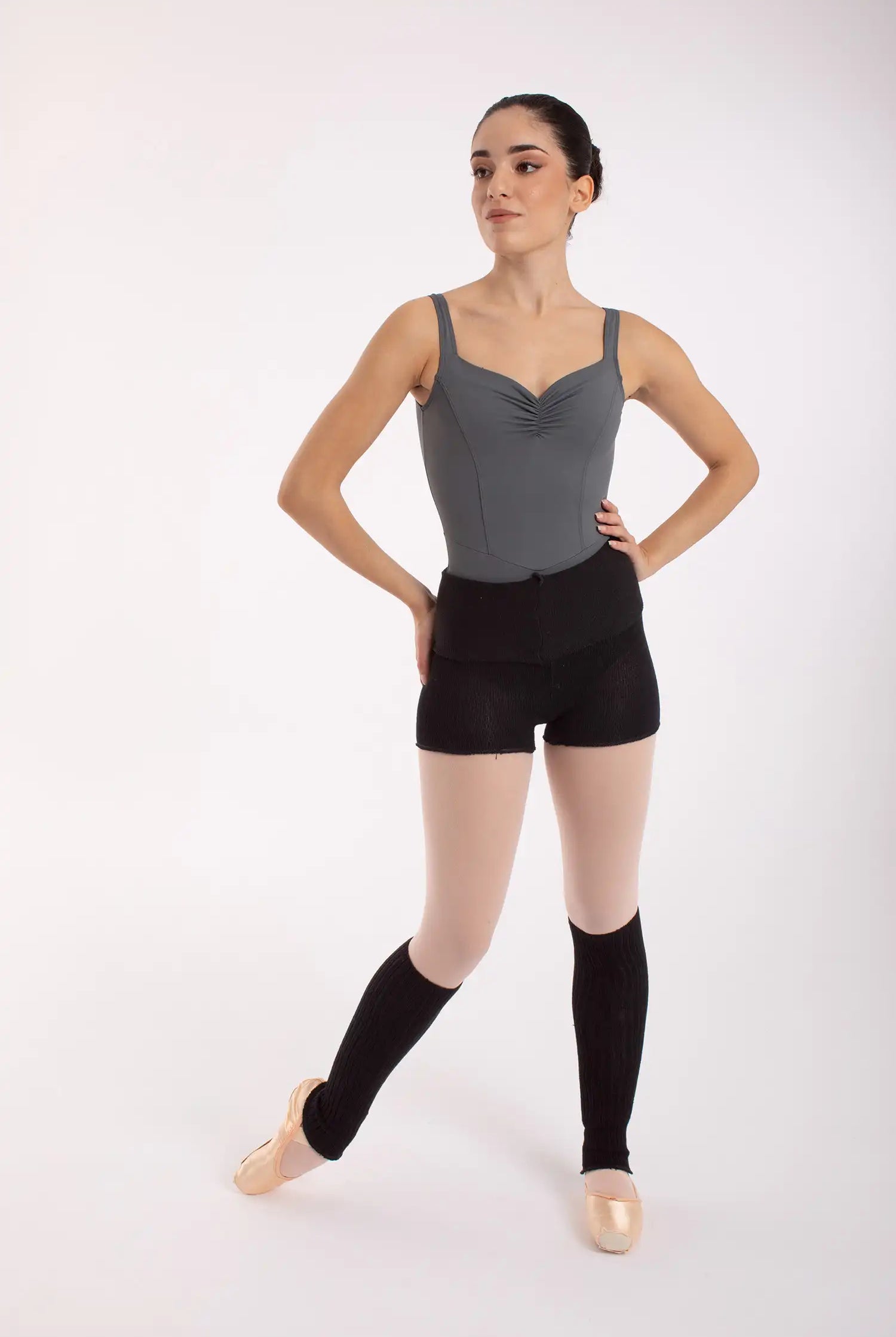 Eine Balletttänzerin, elegant gekleidet in einem grauen Trikot, schwarzen Shorts und 50 cm langen Beinwärmern „Stulpen“ von Intermezzo (Modellnummer 2034 Danae), steht auf Spitzenschuhen und blickt zur Seite vor einem schlichten weißen Hintergrund.