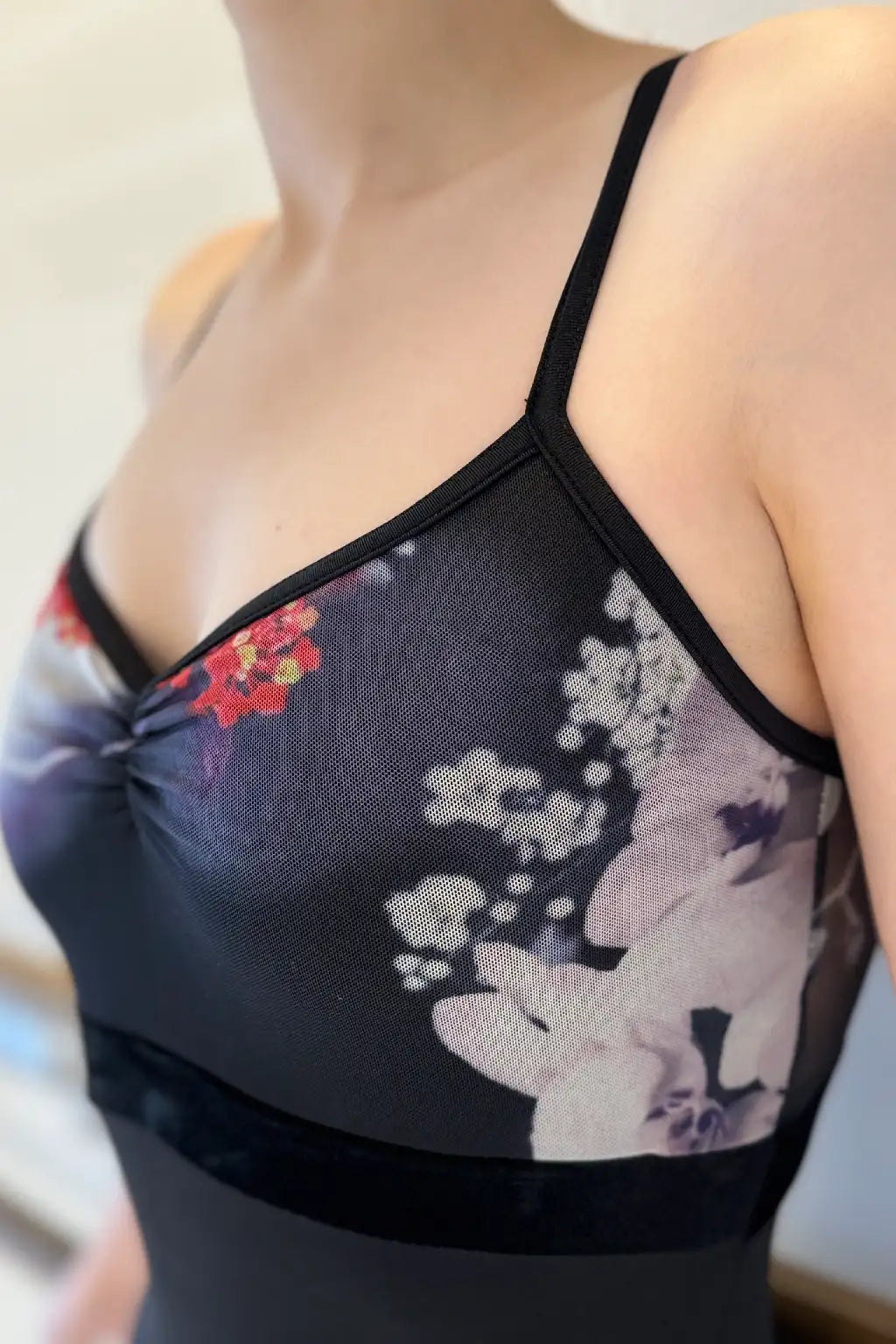 Die Person trägt den Ballettanzug Damen Sansha Gillian 0068M von Dance Line, der ein ärmelloses Oberteil mit Blumenmuster und dünnen Trägern zeigt. Der Stoff zeigt elegant rote und weiße Blumen vor einem sanft verschwommenen Hintergrund.