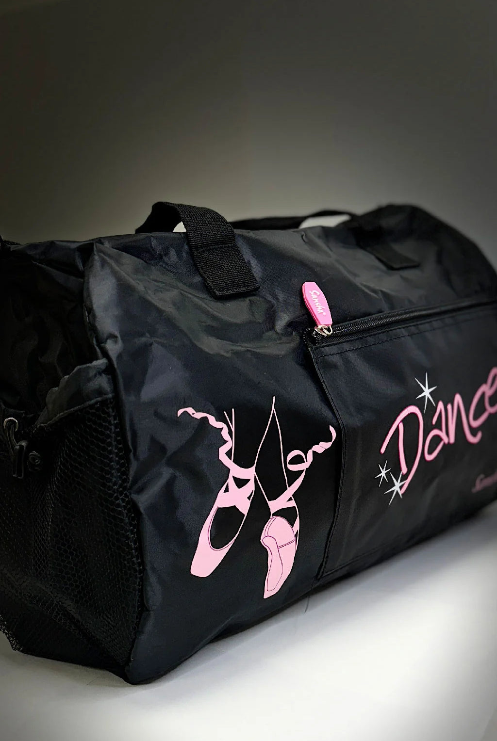 Die Balletttasche Sansha KBAG2 von Dance Line ist eine schwarze Tanztasche mit rosa Ballettschuhen und dem Wort „Dance“ auf der Seite. Sie verfügt über einen rosa Reißverschluss und schwarze Griffe, alles vor einem neutralen Hintergrund.