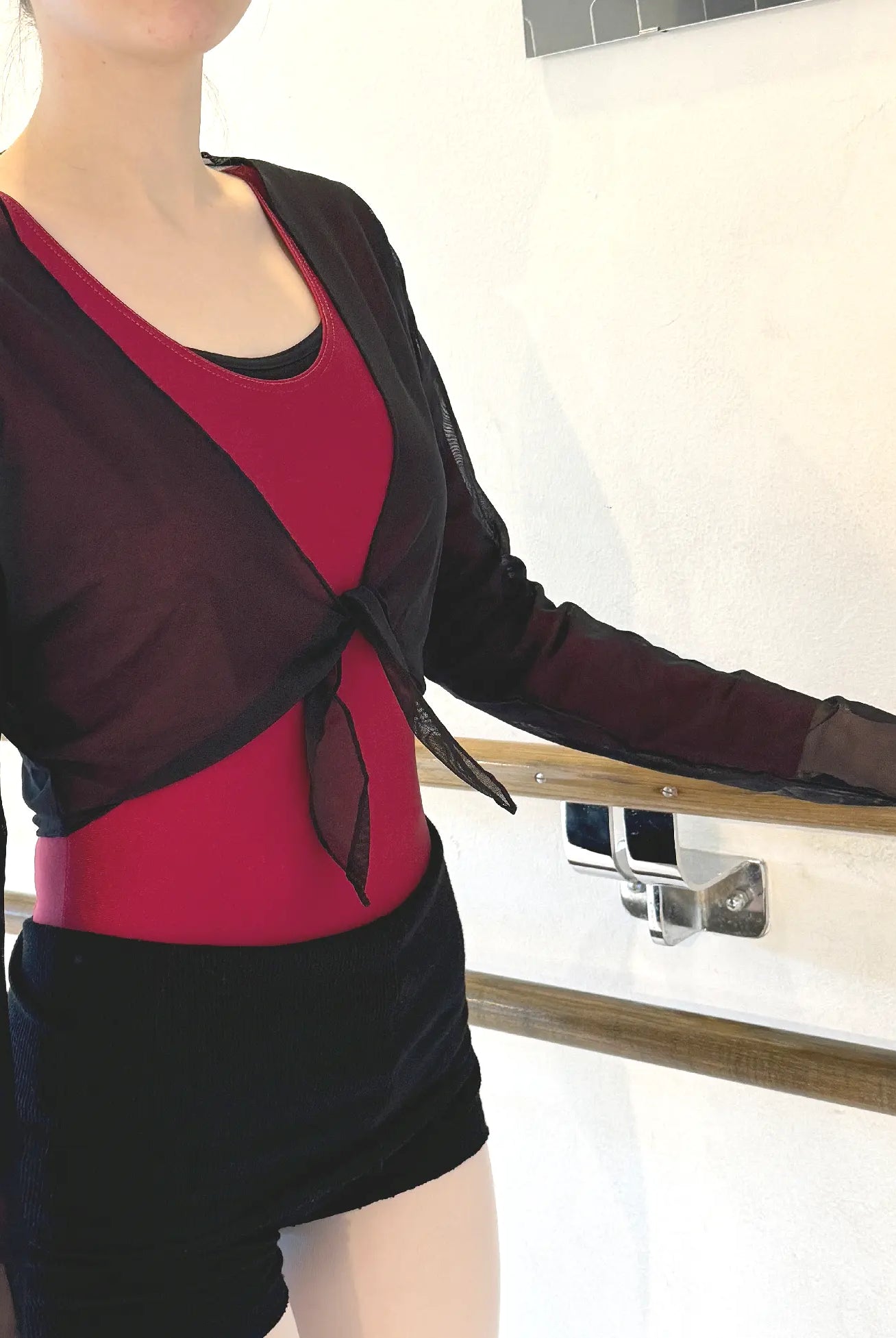 Eine Person, die einen roten Turnanzug, schwarze Shorts und eine Netz-Wickeljacke SoDanca 8175 von SoDanca trägt, steht neben einer Ballettstange vor einer hellen Wand.