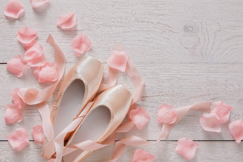 Ein Paar neue Spitzenschuhe mit Bänder liegen auf einem hellen Boden zwischen luftig rosa Blütenblätter.