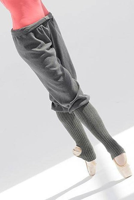 Eine Balletttänzerin in einem rosa Oberteil und grauen Hosen balanciert auf Spitze. Die Füße der Tänzerin stecken in Ballettschuhen mit Beinwärmern der Wärmehose Intermezzo 5076 Rafiness von Intermezzo und liegen auf einer weißen Oberfläche, die bei sanftem Licht einen Schatten wirft.