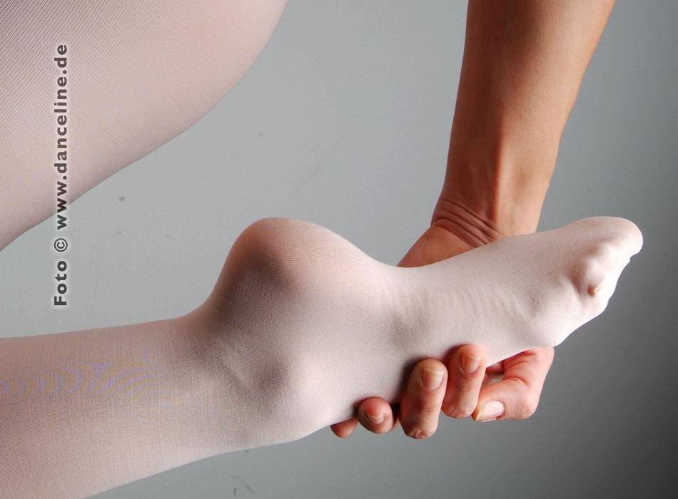 Eine Nahaufnahme des ausgestreckten und gestreckten Fußes einer Person, bekleidet mit einer weißen Ballettstrumpfhose Capezio 1818 B-Tight von Capezio. Die Hand einer anderen Person stützt die Ferse. Der Hintergrund ist schlicht hellgrau. Das Bild weist auf einer Seite ein vertikales Textwasserzeichen „Foto © www.danceline.de“ auf.