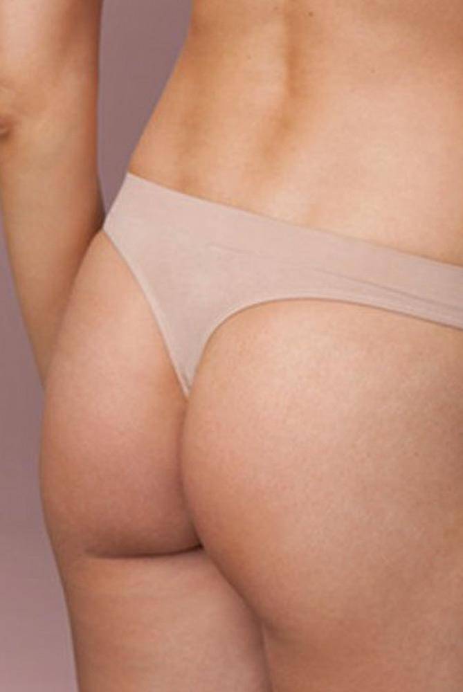 Das Bild zeigt eine Nahaufnahme des unteren Rückens und des Gesäßes einer Person, die einen beigen Capezio Tanga Capezio 3678 Kaya trägt. Der Hintergrund ist mit einem rosa-braunen Farbton verschwommen. Die Haut erscheint glatt und die Person steht mit dem Rücken teilweise zur Kamera, was einen eleganten und doch subtilen Effekt erzeugt.