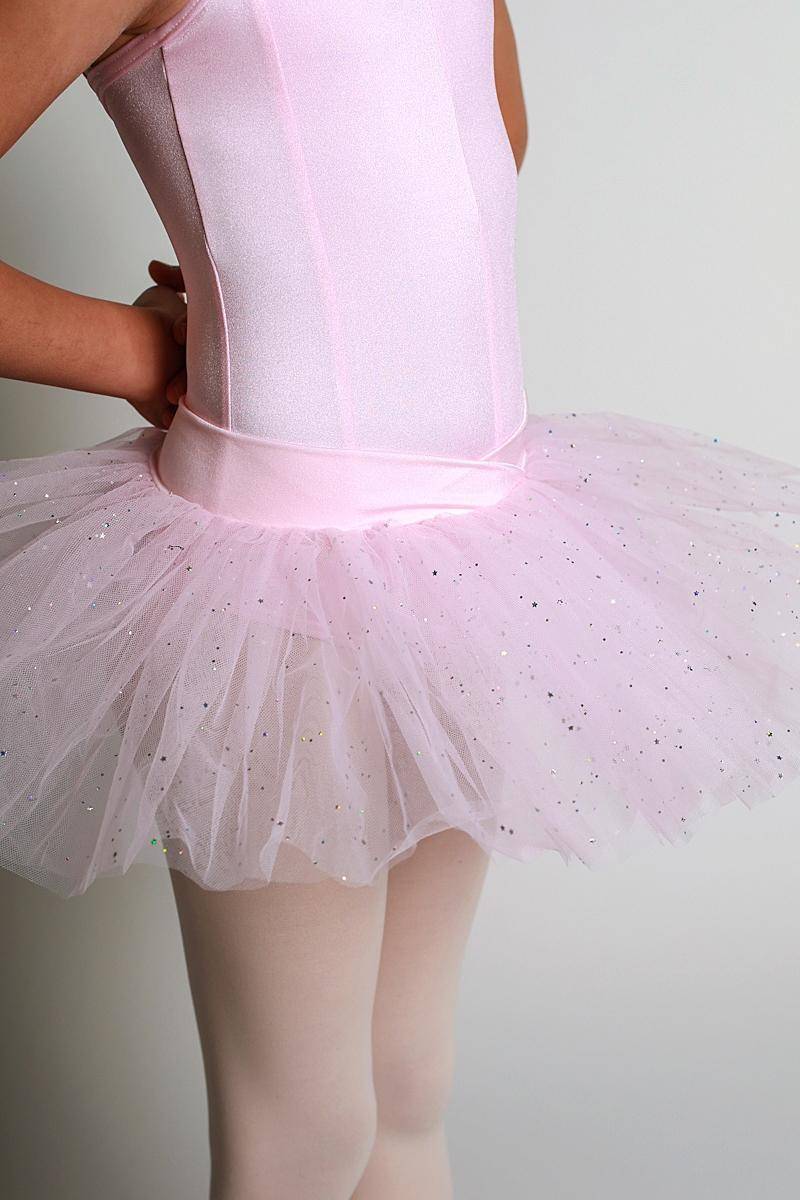 Ein Kind trägt ein rosa Tutulette Capezio 9815C Jada mit Glitzersteinchen und einer weichen Tüllauflage und steht vor einem schlichten Hintergrund, die Hände in die Hüften gestemmt. Der Oberkörper ist sichtbar, wobei das glitzernde Tutu und die weißen Strumpfhosen den Hauptfokus bilden.