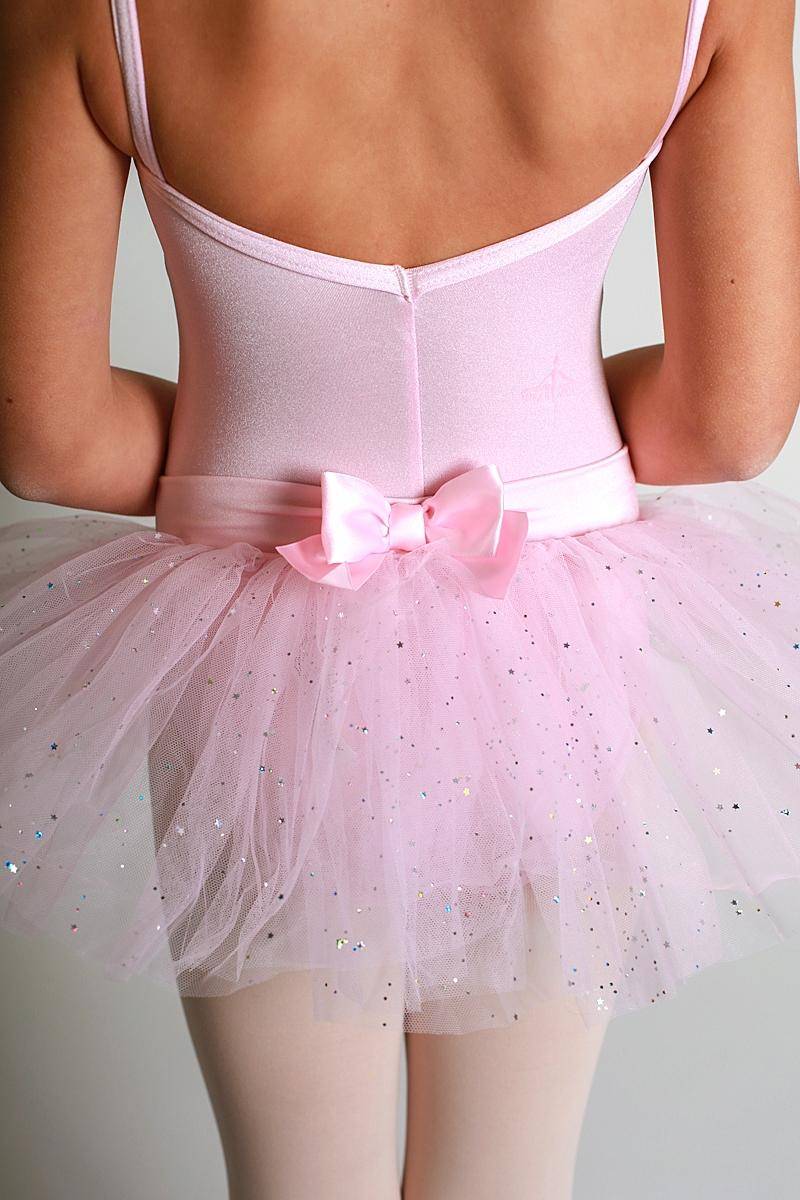 Eine junge Ballerina trägt einen hellrosa Leotard mit dünnen Trägern und eine glitzernd rosa Tutulette Capezio 9815C Jada von Capezio, geschmückt mit einer rosa Satinschleife an der Taille. Sie steht mit dem Rücken zur Kamera und präsentiert das zarte Ensemble aus weichem Tüll und Glitzersteinchen.