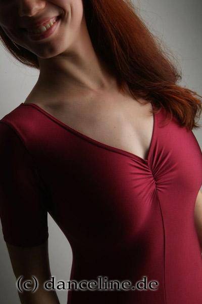 Eine Person mit langen roten Haaren lächelt, während sie den Ganzanzug Danceries G20L Marcea EL von Danceries trägt, der kastanienbraun ist, einen gerafften Ausschnitt und kurze Ärmel hat. Das Foto ist so zugeschnitten, dass nur der Oberkörper und der untere Teil des Gesichts zu sehen sind. Der Hintergrund ist grau und unten ist „danceline.de“ zu sehen.